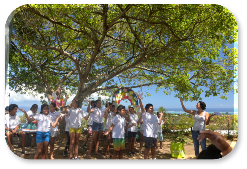Enfants et animatrice du programme Les Arbres du Bonheur dansent avec joie sous un grand arbre lors d'un rituel collectif pour cultiver le bonheur au quotidien
